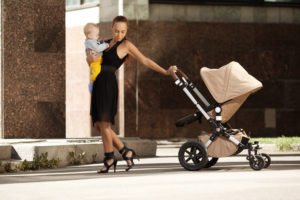 Stylish mom in heels and a dress carrying a baby and holding a stroller after a mommy makeover