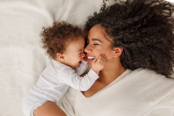 A young mother lies in bed and her infant reaches up to grab her face