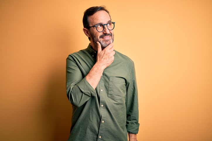 Middle age man wearing casual green shirt and glasses over isolated yellow background with hand on chin thinking about question, pensive expression. Smiling with thoughtful face