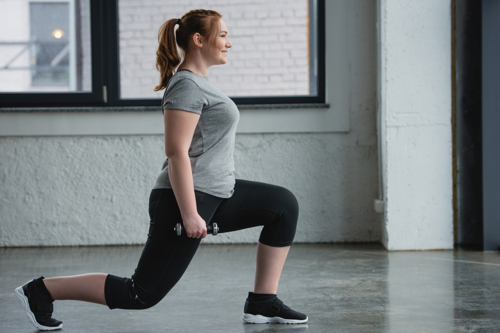 A woman in a workout room wearing fitness clothes does a leg lunge with hand weights