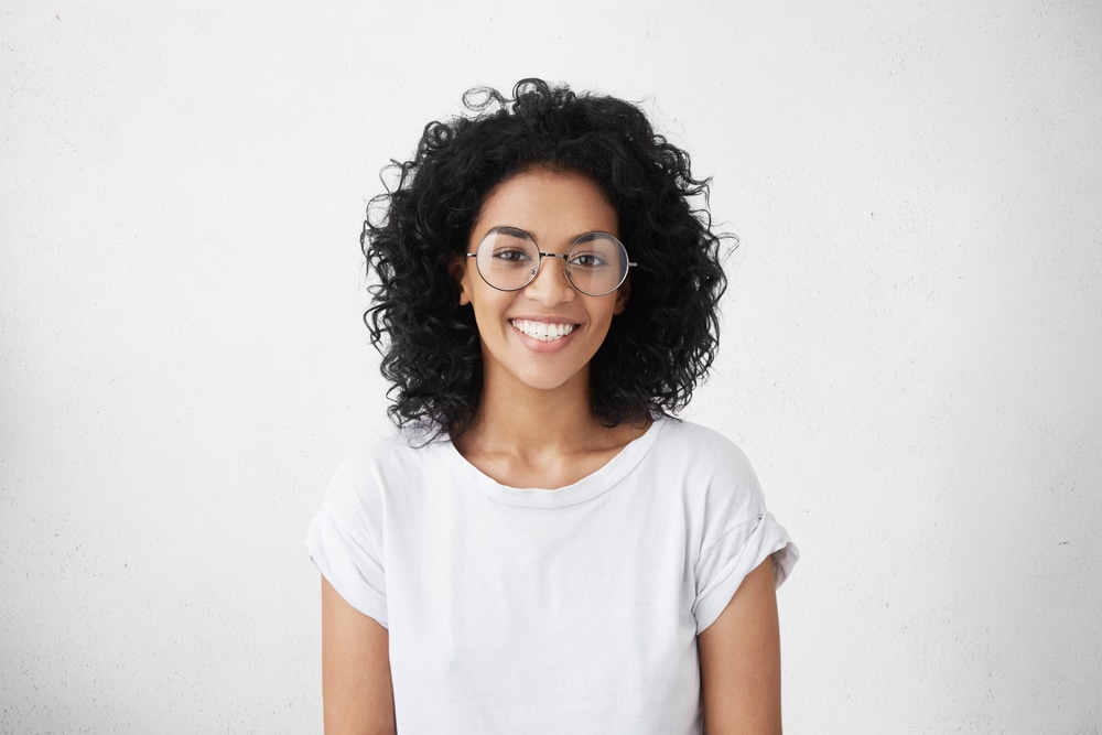 A young woman in a white t-shirt and big round glasses smiles and poses in front of a gray background