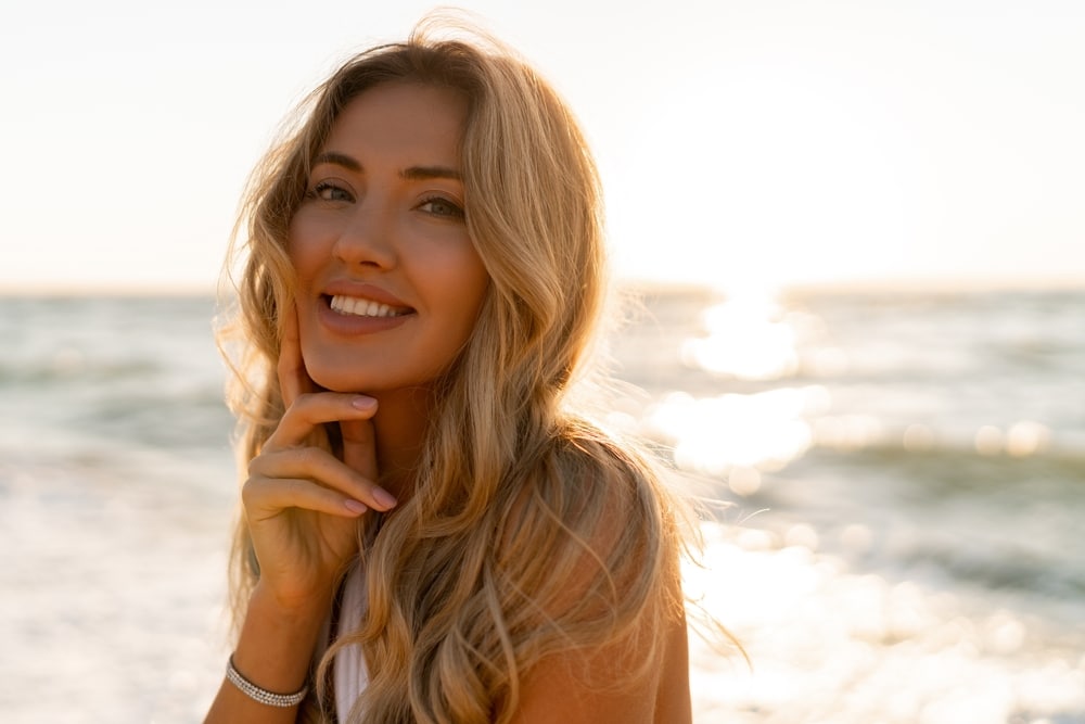 A young woman with wavy hair smiles with her hand to her chin standing in front of the ocean at sunset