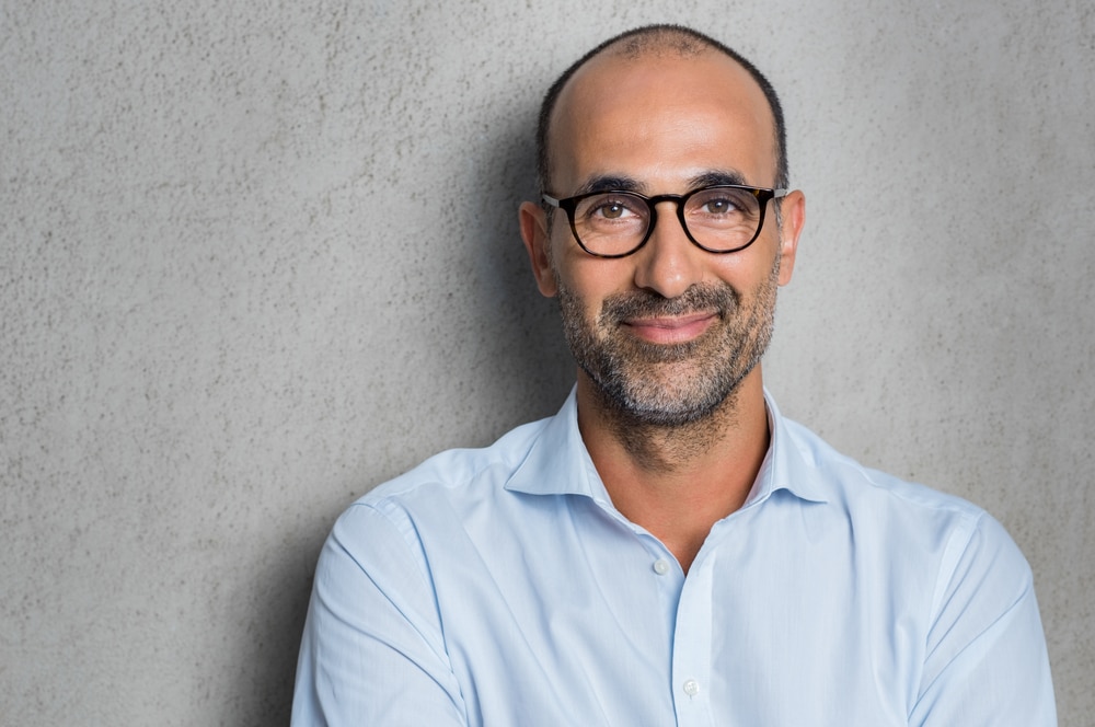 A middle age man smiles slightly at the camera wearing black glasses frames and a button up shirt