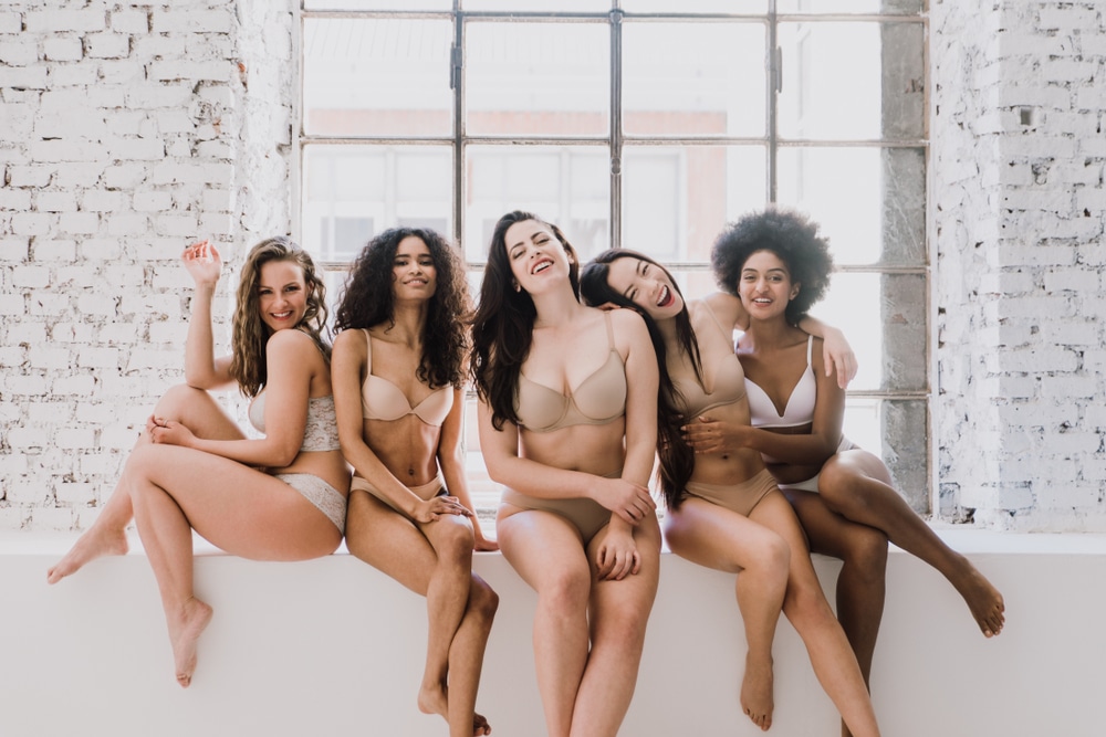 A group of women in bras and panties smile and laugh while seated in front of a large window in a white brick room