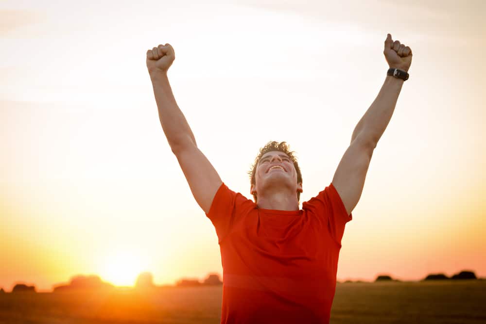 A man in exercise clothes raises his fists in the air triumphantly in front of the rising sun